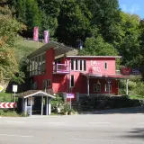 Rotes Haus mit der Aufschrift "Eins + Alles" am Waldrand. Ein kleiner Bushäuschen steht daneben.