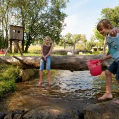 Kinder planschen im Bach auf einem naturnahen Spielplatz. Ein Junge hält einen Eimer, ein Mädchen sitzt auf einem Baumstamm.