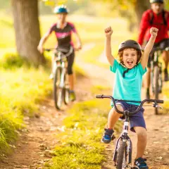 Glückliches Kind auf Fahrrad, feiert Erfolg bei leichten Radtouren mit Kindern. Eltern fahren im Hintergrund.