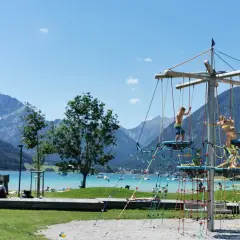 Kinder klettern auf einem Spielgerüst am See. Ein toller Wasserspielplatz für Kinder mit Bergkulisse.
