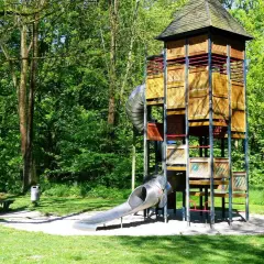 Ein hoher Holzturm mit Rutschen bildet das Zentrum eines abenteuerspielplatzes in Stuttgart. Umgeben von grünen Bäumen lädt er zum Spielen ein.