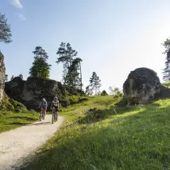 Zwei Radfahrer genießen eine Tour durch eine felsige Landschaft. Sie fahren auf einem Schotterweg, umgeben von grünen Wiesen und Bäumen.