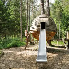 Ein Mädchen klettert auf einem Baumstamm an einem Waldspielplatz. Eine runde Holzhütte mit Rutsche steht im Mittelpunkt.