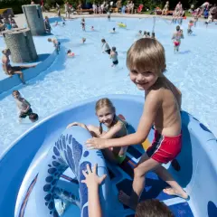 Fröhliche Kinder planschen und rutschen im Freibad. Ein Junge grinst in die Kamera, während ein Mädchen auf der blauen Rutsche sitzt. Ein perfekter Tag im bayerischen Wald mit Kindern.