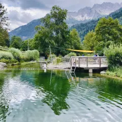 Ein idyllischer Badesee in Bad Hindelang, perfekt für das Wandern mit Kindern. Ein Steg mit Leiter lädt zum Sprung ins kühle Nass ein, während im Hintergrund majestätische Berge thronen.