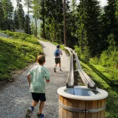 Zwei Kinder rennen auf einem Schotterweg an einer **outdoor kugelbahn** vorbei, umgeben von einem Wald und grünen Hängen.