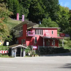 Rotes Haus mit der Aufschrift "Eins + Alles" am Waldrand. Ein kleiner Bushäuschen steht daneben.