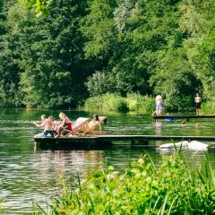 Menschen entspannen sich auf einem Floß und genießen die Sonne an einem Münchner See. Andere stehen auf einem Steg, einige baden im Wasser.