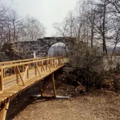 Holzbrücke führt zu einer steinernen Torbogenruine von Hofen, umgeben von kahlen Bäumen und Herbstlaub.
