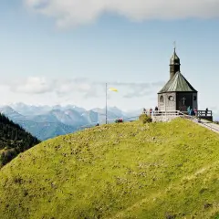 Blick auf das alte Wallberghaus auf einem grünen Hügel mit Bergpanorama. Eine Wanderung hierher lohnt sich für diese Aussicht.