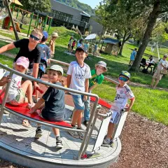 Kinder drehen sich auf einem Karussell auf dem Spielplatz. Ein Junge mit Sonnenbrille lacht, während er sich festhält. Der Spielplatz hat auch eine Schaukel und eine Rutsche, die an einen Flugzeugflügel erinnert.