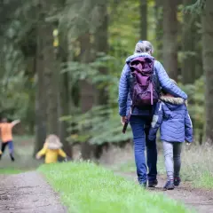 Familie genießt einen Waldspaziergang bei Aschaffenburg. Kinder in gelben Jacken rennen voraus, während eine Frau mit einem Kind Hand in Hand den Weg entlanggeht.