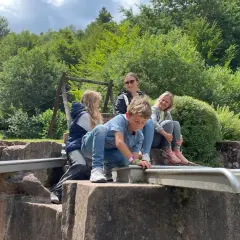 Familie genießt einen sonnigen Tag beim Wandern mit Kindern in der Pfalz. Ein Junge spielt am Wasserlauf, während die anderen entspannt zusehen.