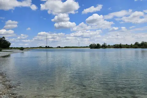 Ein sonniger Tag am Hollerner See mit blauem Himmel, weißen Wolken und klarem Wasser.