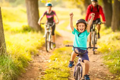 Glückliches Kind auf Fahrrad, feiert Erfolg bei leichten Radtouren mit Kindern. Eltern fahren im Hintergrund.