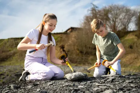 Zwei Kinder knien im Freien und graben mit Hämmern und Meißeln nach Fossilien. Vielleicht finden sie bald etwas Spannendes, wie bei Ausgrabungen in Stuttgart.