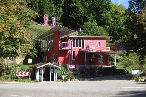 Rotes Haus mit der Aufschrift "Eins + Alles" am Waldrand. Ein kleiner Bushäuschen steht daneben.