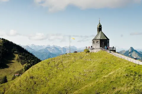 Blick auf das alte Wallberghaus auf einem grünen Hügel mit Bergpanorama. Eine Wanderung hierher lohnt sich für diese Aussicht.