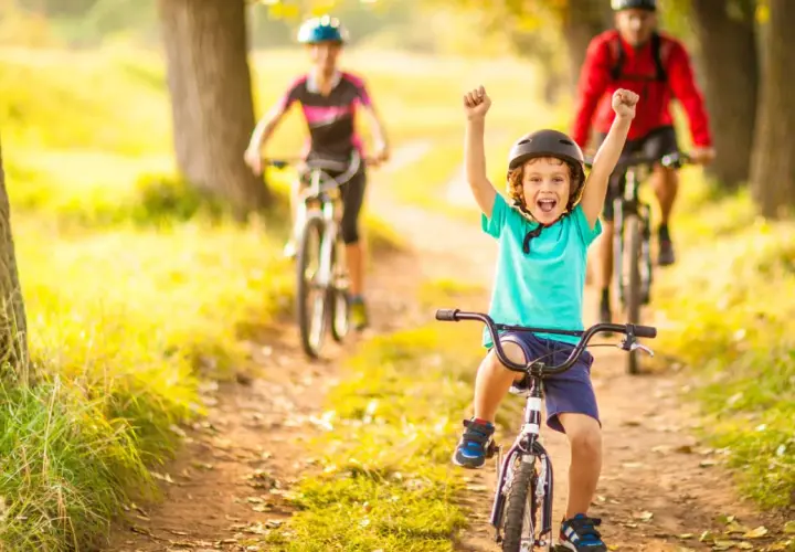Glückliches Kind auf Fahrrad, feiert Erfolg bei leichten Radtouren mit Kindern. Eltern fahren im Hintergrund.