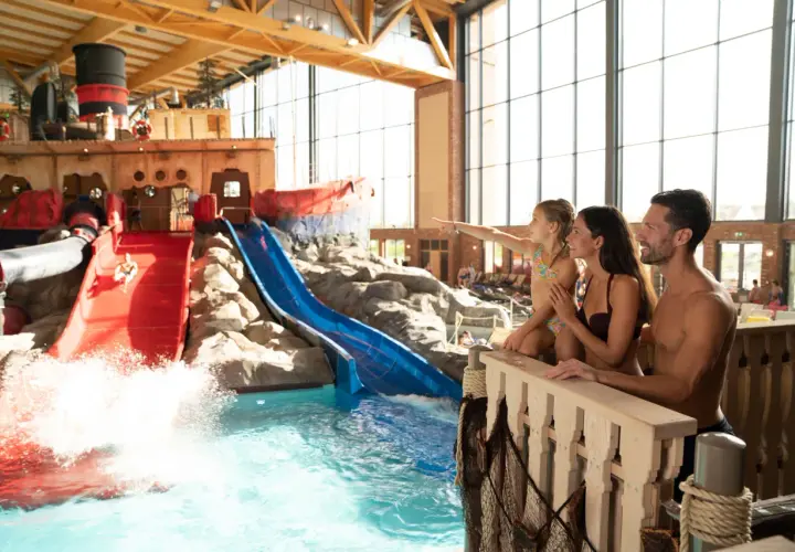 Familie genießt die **Therme mit Kindern**. Ein Junge rutscht auf einer roten Wasserrutsche, während die Familie zusieht.