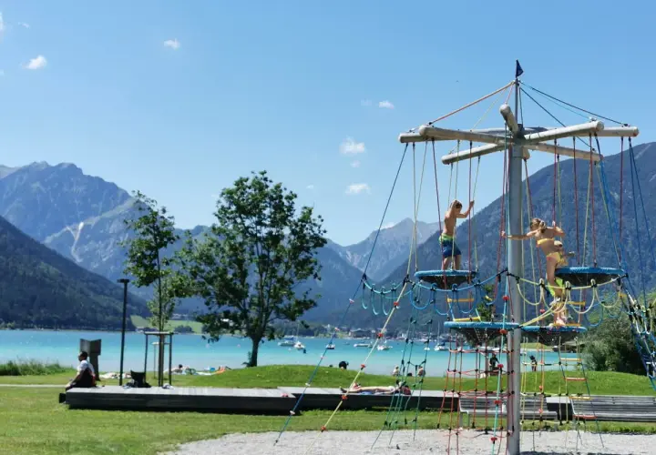 Kinder klettern auf einem Spielgerüst am See. Ein toller Wasserspielplatz für Kinder mit Bergkulisse.