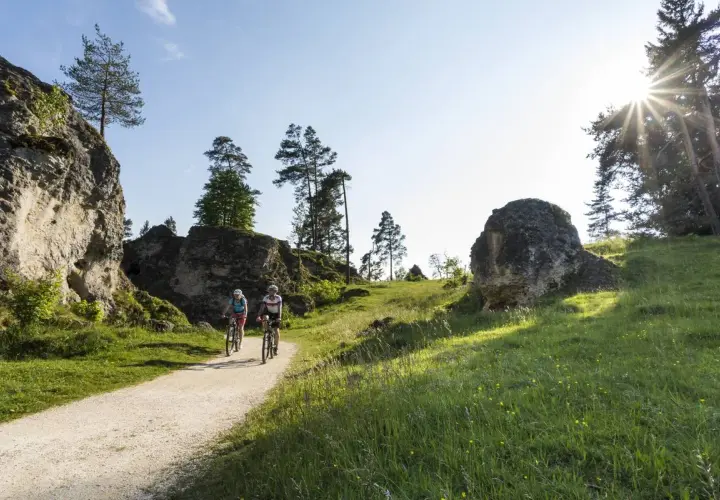 Zwei Radfahrer genießen eine Tour durch eine felsige Landschaft. Sie fahren auf einem Schotterweg, umgeben von grünen Wiesen und Bäumen.