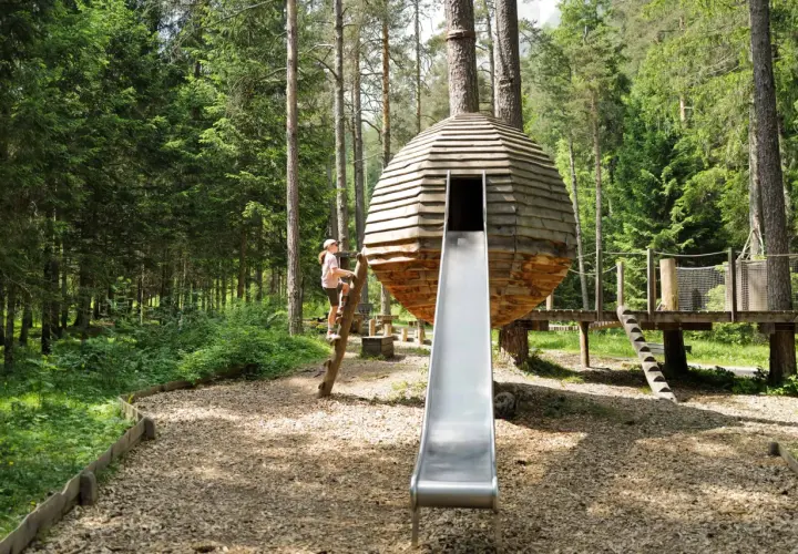 Ein Mädchen klettert auf einem Baumstamm an einem Waldspielplatz. Eine runde Holzhütte mit Rutsche steht im Mittelpunkt.