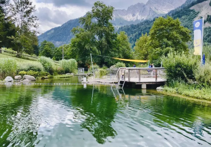 Ein idyllischer Badesee in Bad Hindelang, perfekt für das Wandern mit Kindern. Ein Steg mit Leiter lädt zum Sprung ins kühle Nass ein, während im Hintergrund majestätische Berge thronen.