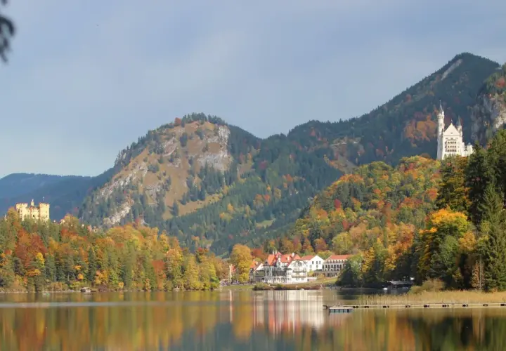Zwei Märchenschlösser, Hohenschwangau und Neuschwanstein, thronen über einem See in Bayern, umgeben von bunten Herbstwäldern.