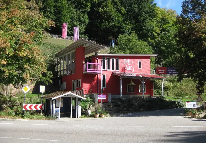 Rotes Haus mit der Aufschrift "Eins + Alles" am Waldrand. Ein kleiner Bushäuschen steht daneben.