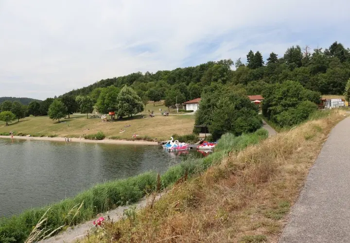 Sonniger Tag am badesee Zaberfeld. Menschen entspannen am Ufer, Kinder planschen im Wasser, Tretboote warten auf Ausflügler.