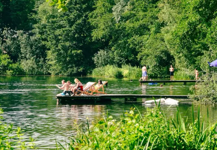 Menschen entspannen sich auf einem Floß und genießen die Sonne an einem Münchner See. Andere stehen auf einem Steg, einige baden im Wasser.