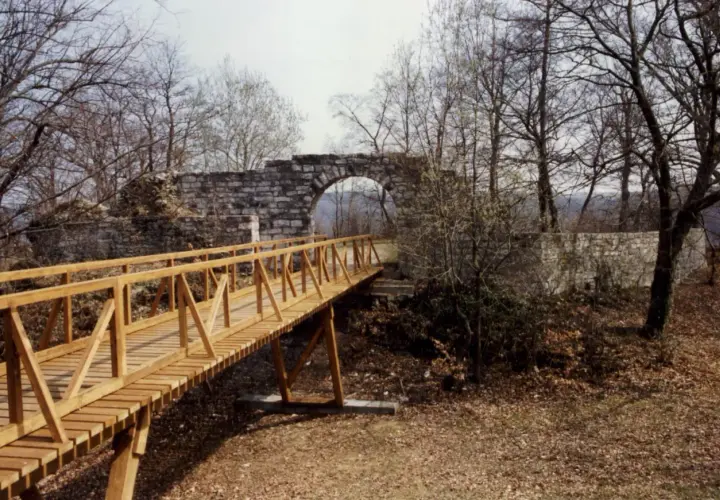 Holzbrücke führt zu einer steinernen Torbogenruine von Hofen, umgeben von kahlen Bäumen und Herbstlaub.