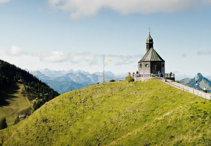 Blick auf das alte Wallberghaus auf einem grünen Hügel mit Bergpanorama. Eine Wanderung hierher lohnt sich für diese Aussicht.