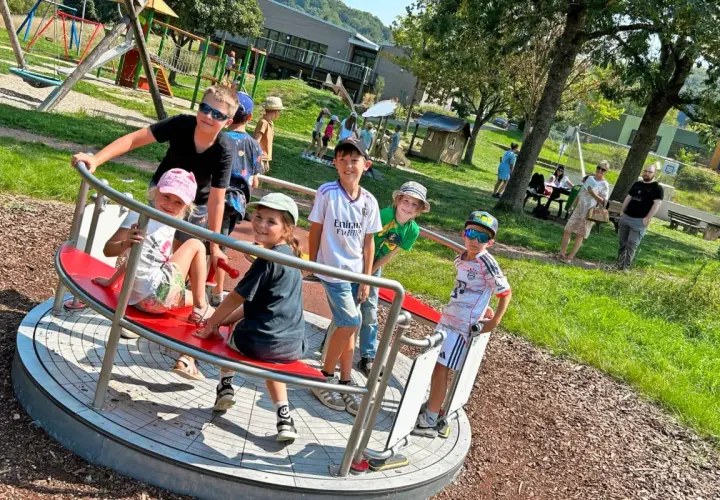 Kinder drehen sich auf einem Karussell auf dem Spielplatz. Ein Junge mit Sonnenbrille lacht, während er sich festhält. Der Spielplatz hat auch eine Schaukel und eine Rutsche, die an einen Flugzeugflügel erinnert.