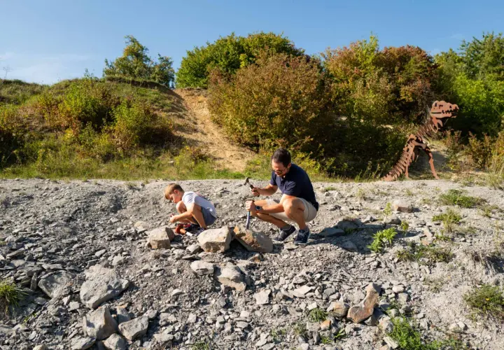 Ein Vater und sein Sohn graben nach Fossilien. Im Hintergrund steht ein Skelett eines Dinosauriers.