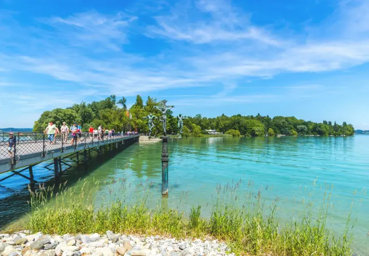 Menschen spazieren auf einer Brücke am Bodensee. Ein Denkmal mit Kreuzigungsgruppe steht im Wasser. Hier kann man in Konstanz viel unternehmen.