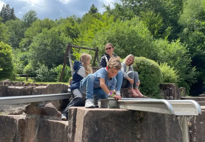 Familie genießt einen sonnigen Tag beim Wandern mit Kindern in der Pfalz. Ein Junge spielt am Wasserlauf, während die anderen entspannt zusehen.