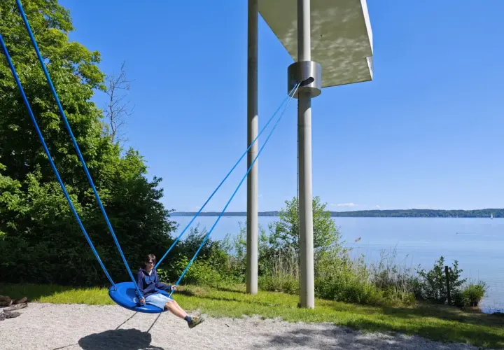 Ein Junge schaukelt am Starnberger See. Kinder genießen die Sonne und den Blick auf das Wasser.