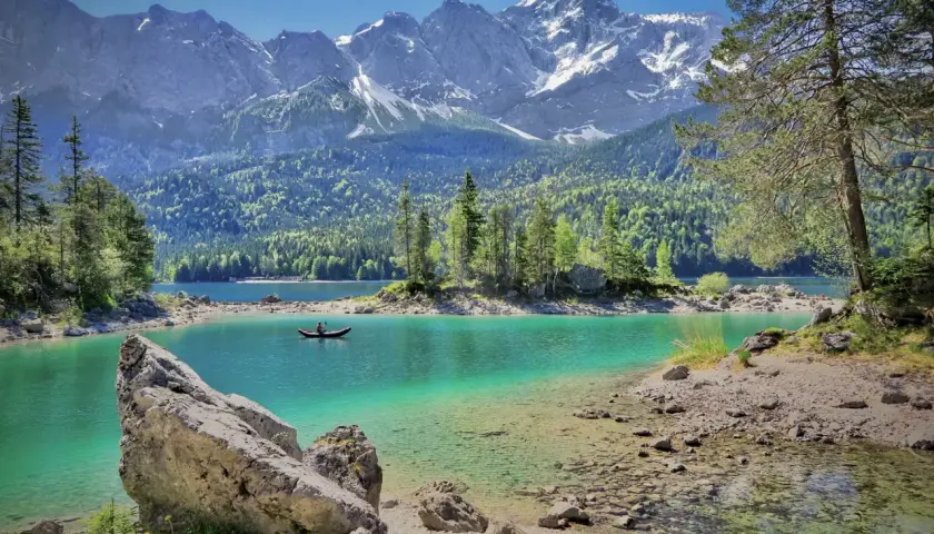 Ein einsamer Kanufahrer auf einem türkisfarbenen See, umgeben von grünen Wäldern und schneebedeckten Bergen. Perfekt für einen Tagesausflug in Bayern.