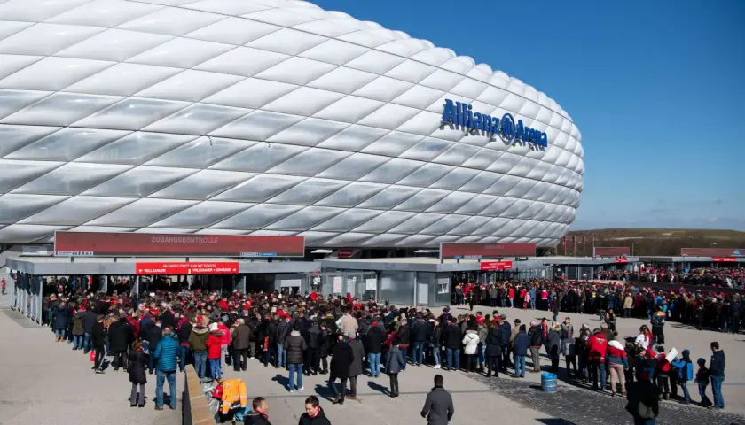 Menschenmassen warten vor der Allianz Arena Spielwelt auf Einlass. Die futuristische Fassade des Stadions leuchtet im Sonnenlicht.