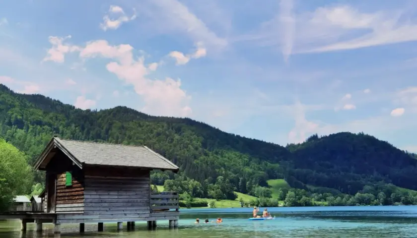 Holzhütte am See, Menschen planschen im Wasser. Perfekt für **Bayrischzell wandern mit Kindern** und Erfrischung.