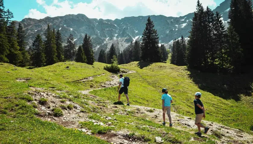 Familie wandert auf einem steinigen Pfad, dem Schmugglerpfad Oberjoch, durch grüne Almwiesen mit Nadelbäumen und schneebedeckten Bergen im Hintergrund.