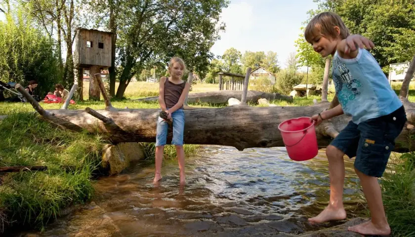 Kinder planschen im Bach auf einem naturnahen Spielplatz. Ein Junge hält einen Eimer, ein Mädchen sitzt auf einem Baumstamm.