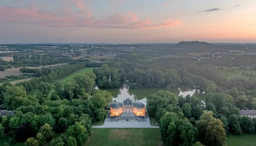 Schloss Monrepos Ludwigsburg, beleuchtet im Abendlicht, umgeben von einem See und üppigen Bäumen, mit Blick auf die Stadt.