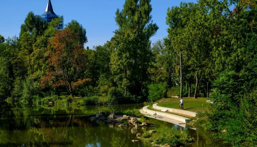 Ein Radfahrer fährt auf einem Weg am Wasser entlang, umgeben von Bäumen und einem Kirchturm.