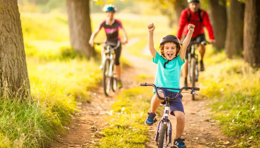Glückliches Kind auf Fahrrad, feiert Erfolg bei leichten Radtouren mit Kindern. Eltern fahren im Hintergrund.