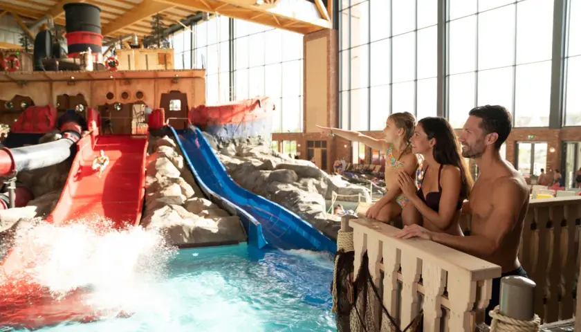 Familie genießt die **Therme mit Kindern**. Ein Junge rutscht auf einer roten Wasserrutsche, während die Familie zusieht.