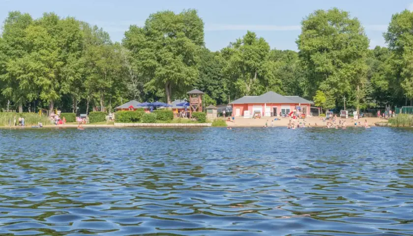 Sommerliche Szene an einem der **Badeseen Berlin** mit vielen Menschen am Strand und im Wasser, umgeben von grünen Bäumen.