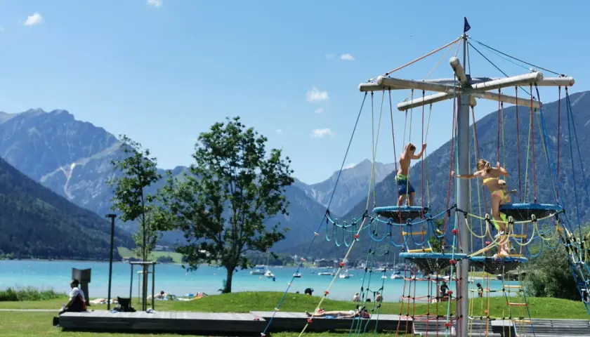 Kinder klettern auf einem Spielgerüst am See. Ein toller Wasserspielplatz für Kinder mit Bergkulisse.